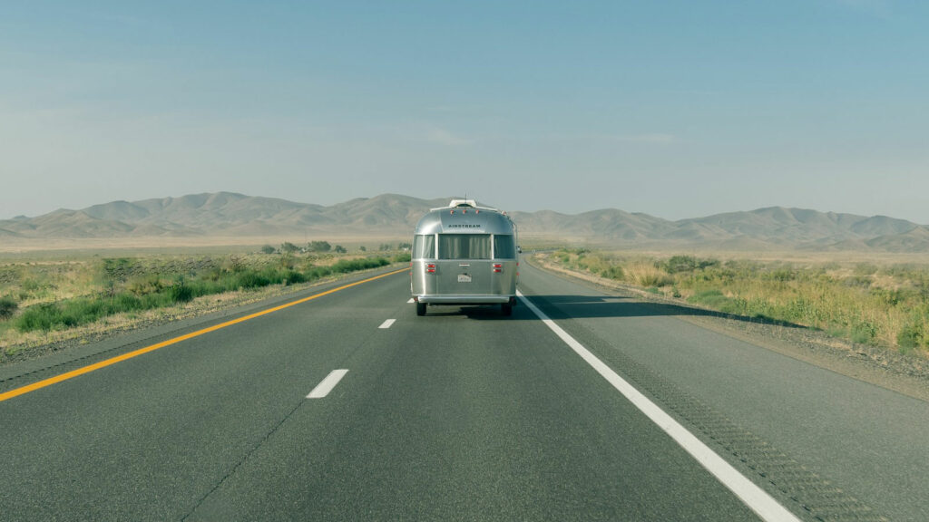 A shiny silver Airstream travel trailer heading down a two lane highway towards mountains in the distance.