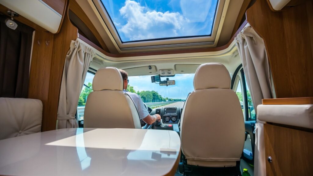 A view of a driver in the pilot seat of a motorhome cruising down the highway as the sun shines in through the sun roof above.