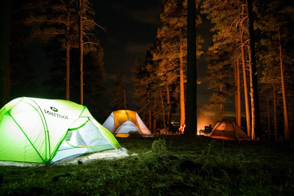 Green and orange tents lit up with flashlights and a campfire burning in the background while campers enjoy the night.