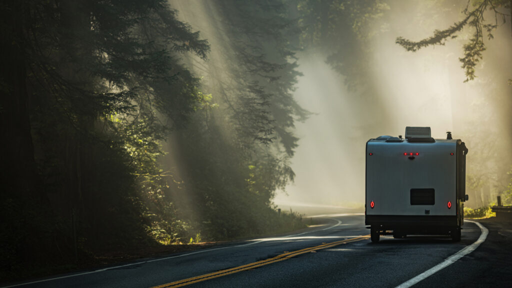 A travel trailer's brake lights are glowing red as the driver maneuvers through a foggy windy forest road.
