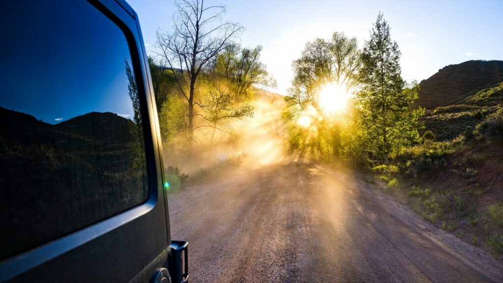 A van drives down a dirt road and kicks up dust that the setting sun filters through.