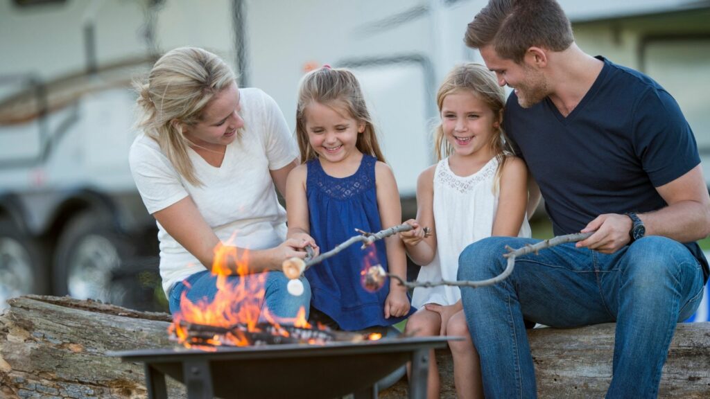 A family of four roasts marshmallows on sticks over a portable fire pit with their RV in the background.