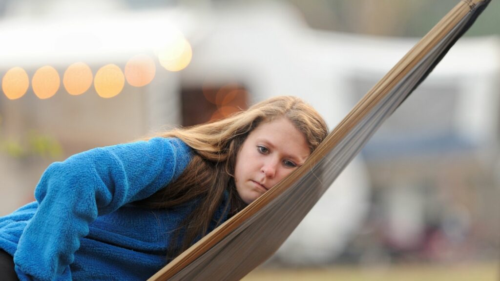 An adolescent girl looks sad and bored sitting in a hammock at an RV park.
