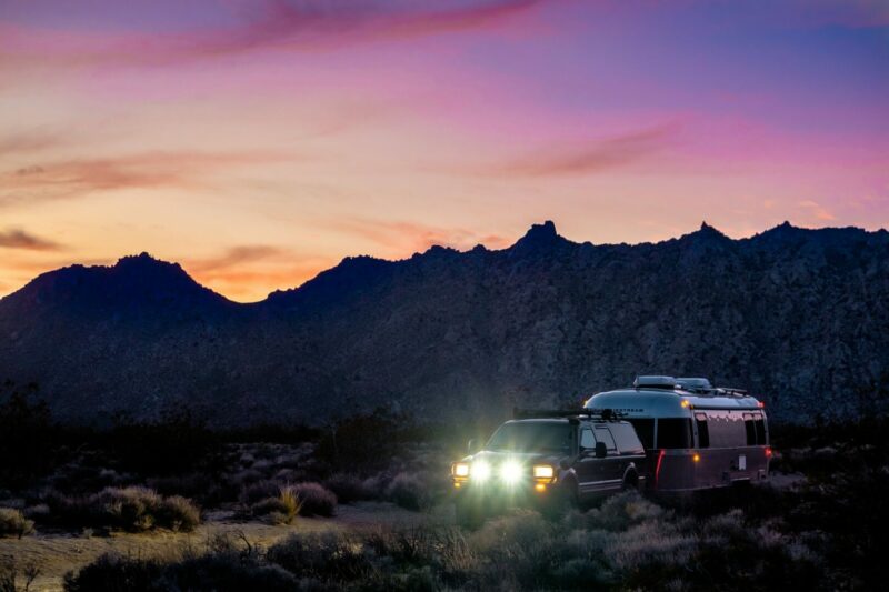 Sun sets of cliffs in Arizona, coloring the sky in yellow, pink, and blue as a travel trailer makes it way down a dirt road.