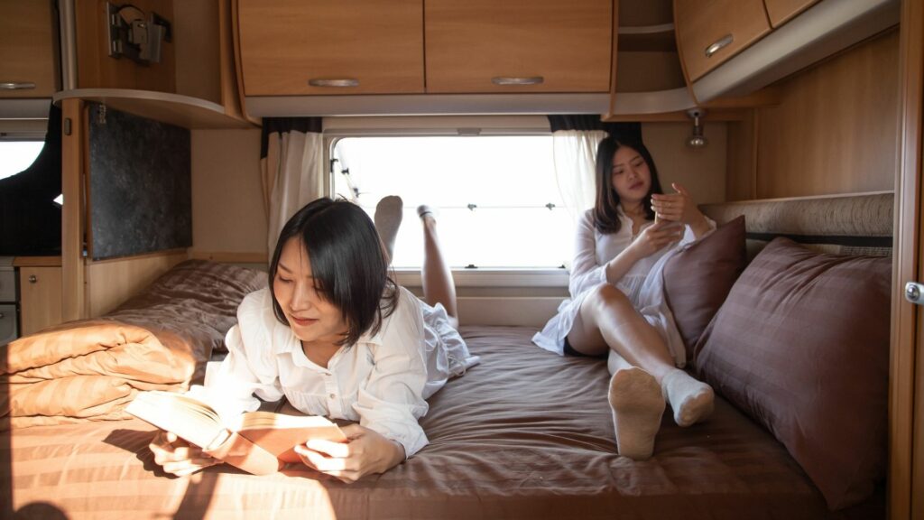 Two women relax by reading and using their phones on the RV mattress in their motorhome.