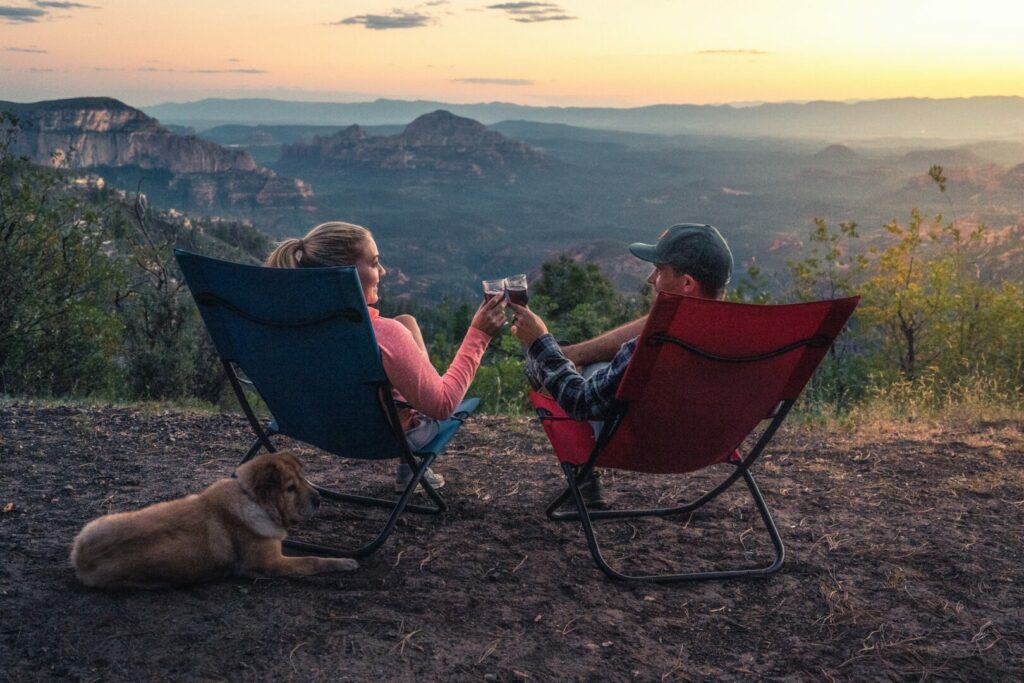 Two campers cheers and enjoy a glass of wine in their camping chairs overlooking a beautiful sunset over the mountains with their dog.