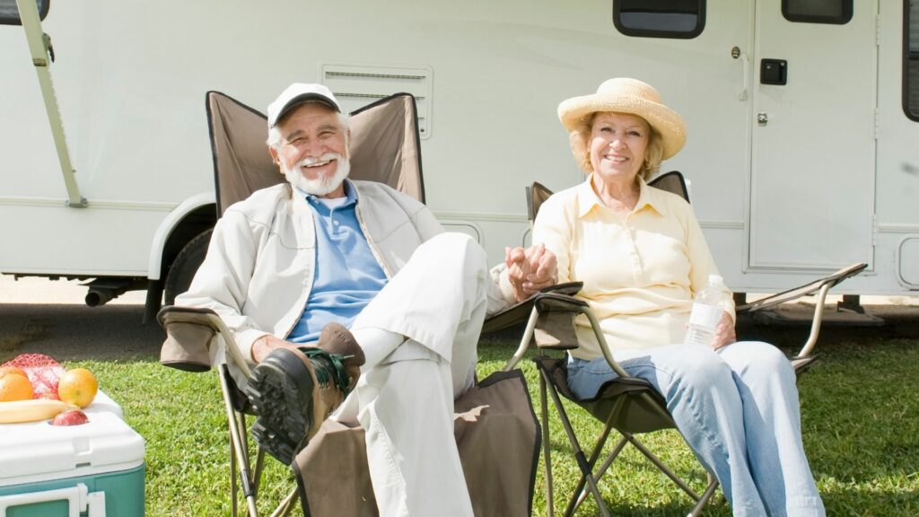 Senior couple sitting outside an RV in the sunshine with hats on, holding hands.