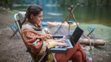 Cheerful woman uses her computer to plan the rest of her trip while sitting in a camp chair next to the fire while camping on a river.
