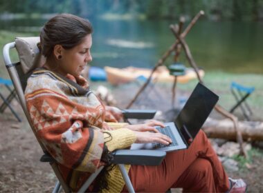 Cheerful woman uses her computer to plan the rest of her trip while sitting in a camp chair next to the fire while camping on a river.