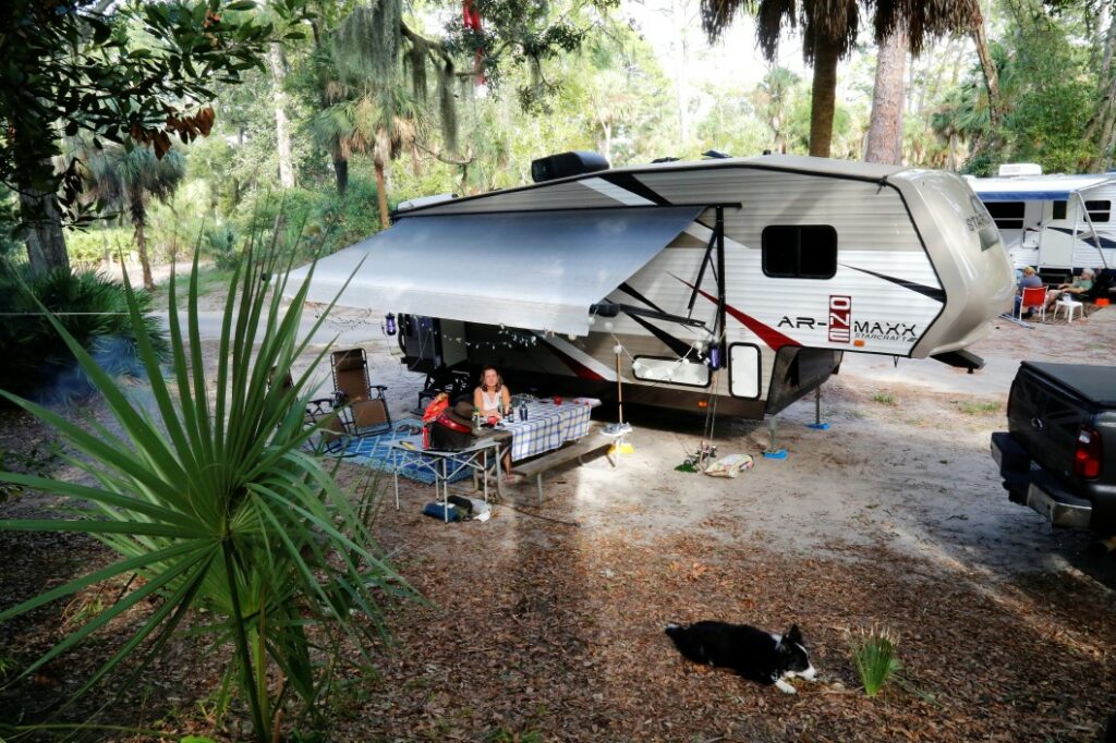 A woman sits outside of her fifth wheel in a tropical campsite.