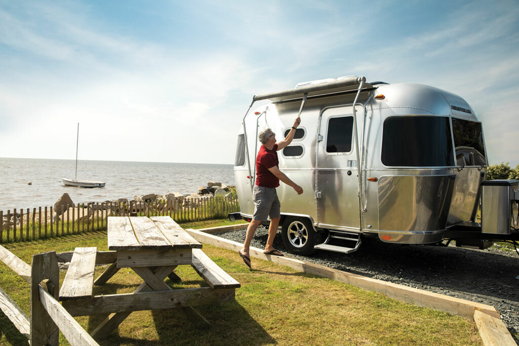 A lifestyle photo of a man pulling out the awning on a Caravel travel trailer parked in a campsite near the ocean.