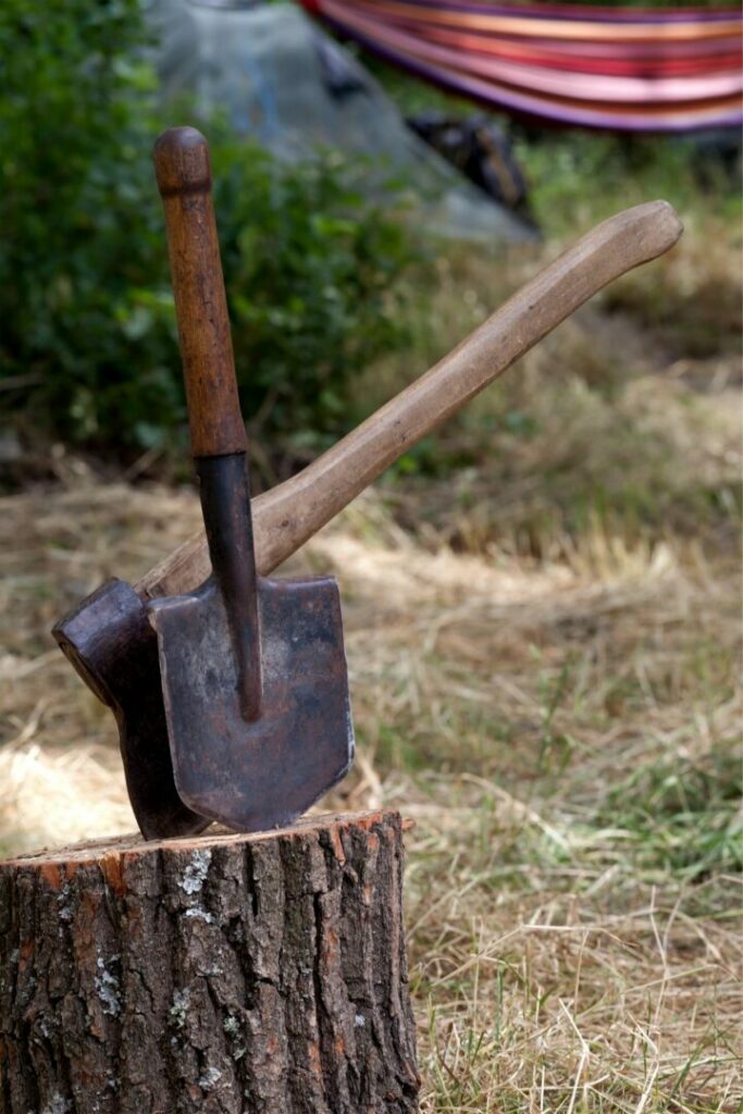 Shovel and axe stuck into a tree stump out in the woods with a hammock in the background.