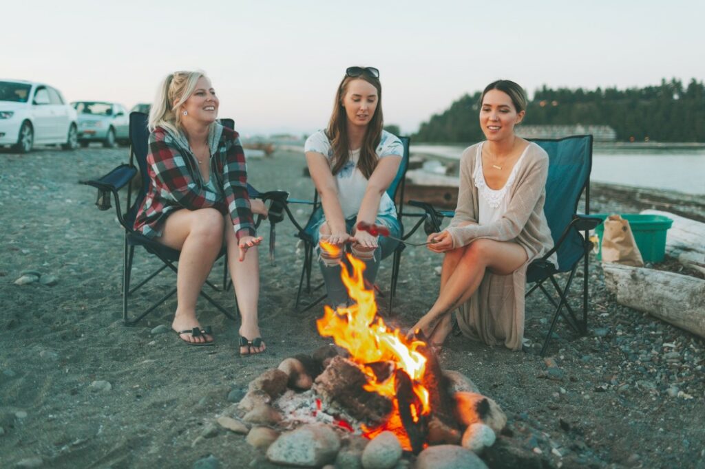 Three woman camping on the beach cook hotdogs over the fire with campfire roasting sticks