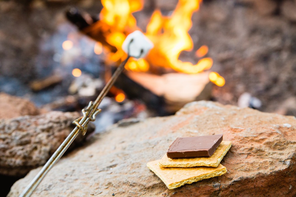 A metal campfire roasting stick cooks a marshmallow over a fire and graham crackers and chocolate are ready to be made into a smore.
