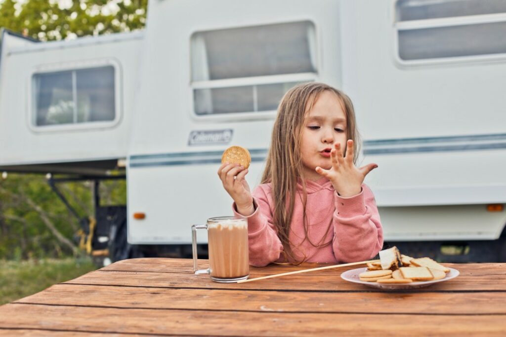 A young girl enjoys cookies, hot cocoa, and a roasted marshmallow at a picnic table outside an RV.