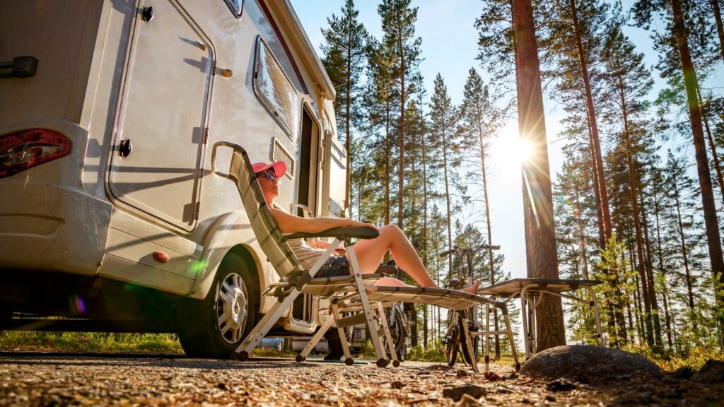 A woman reclines in the sun outside of her motorhome in the woods.