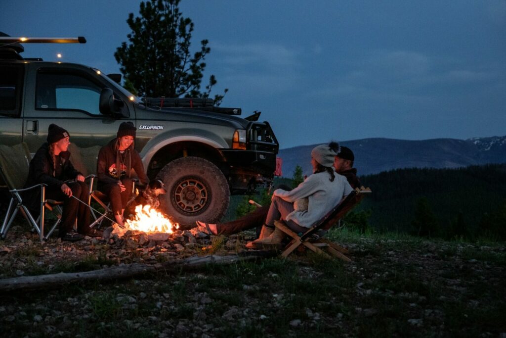 Friends sit around the campfire at dusk beside their off-road vehicle