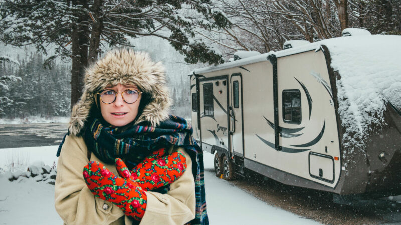 A woma bundled in mittens, fur hat, coat, and scarf in the winter outside her snow covered RV