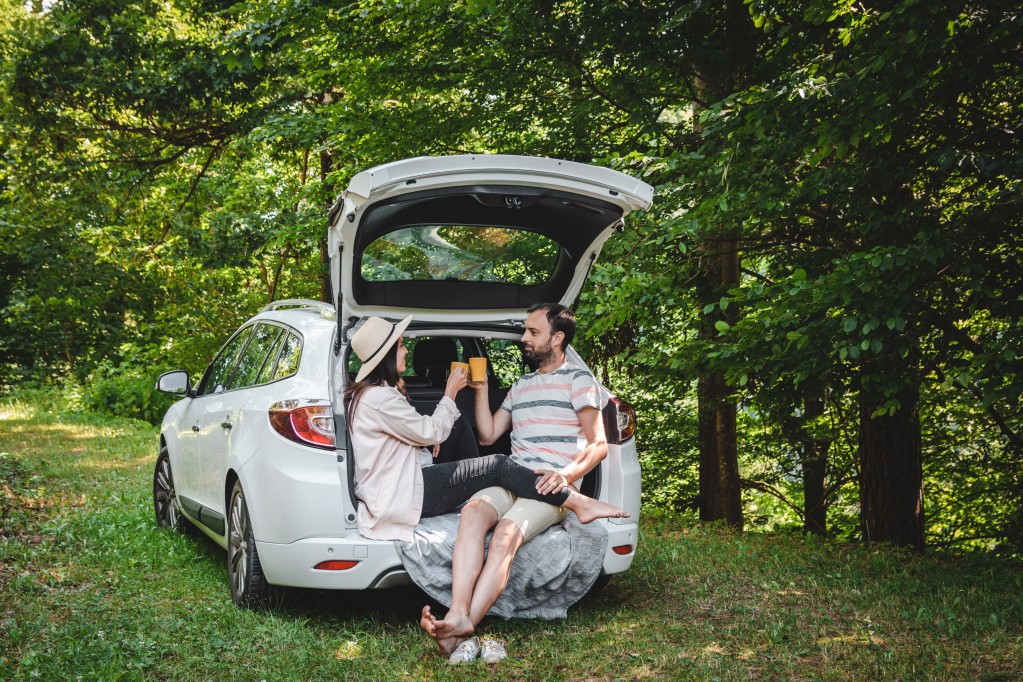 A couple sits in the trunk of their car enjoying a beverage while camping out in the woods