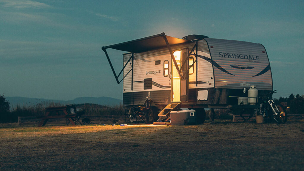 A travel trailer camping in nature, inside lights shine out the front door at dusk.
