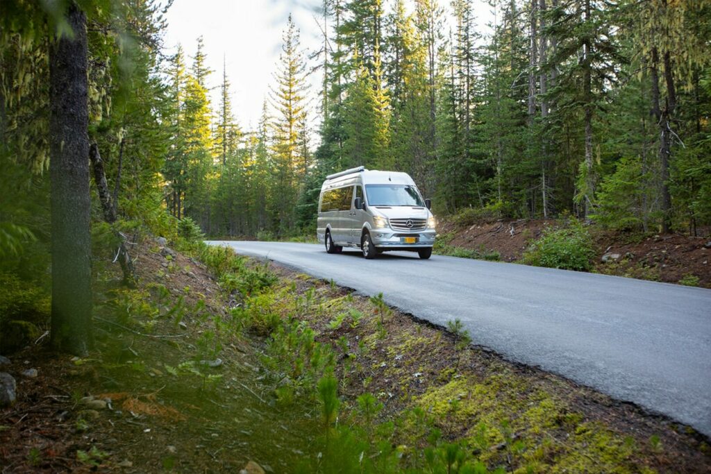 A luxury Class B travel van cruises down a paved road through the wilderness.