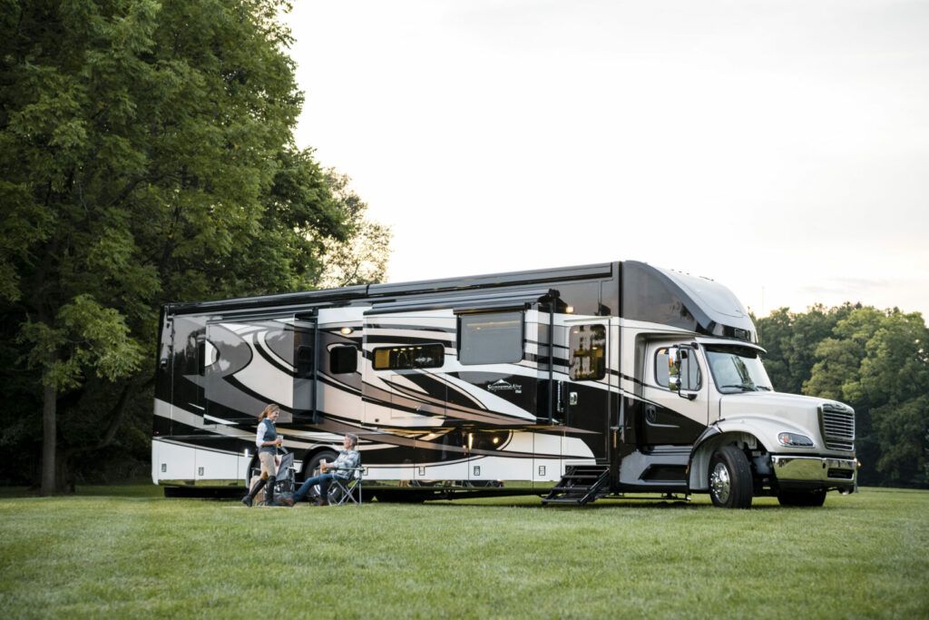 A couple sits outside their luxury Newmar coach in a lush grassy field surrounding by trees.