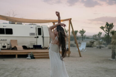 A graceful woman hold her arms above her head in a dress outside of a luxury travel trailer in the desert.