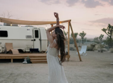 A graceful woman hold her arms above her head in a dress outside of a luxury travel trailer in the desert.