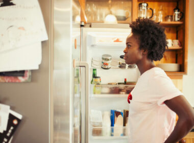 A woman with dark hair stand in front of her fridge with the door open, looking inside and thinking.