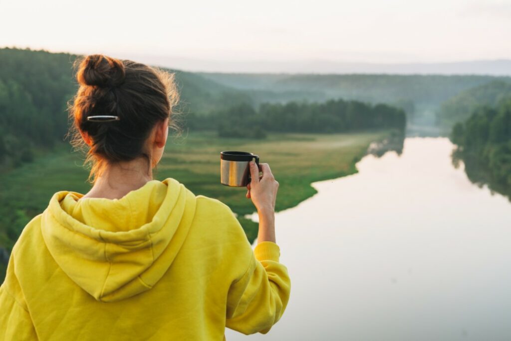 A woman in a hoodie holds her coffee mug out as she overlooks a river and meadow from her campsite