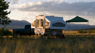 A pop up camper has made a nice campsite out in an open field with some trees and mountains nearby.