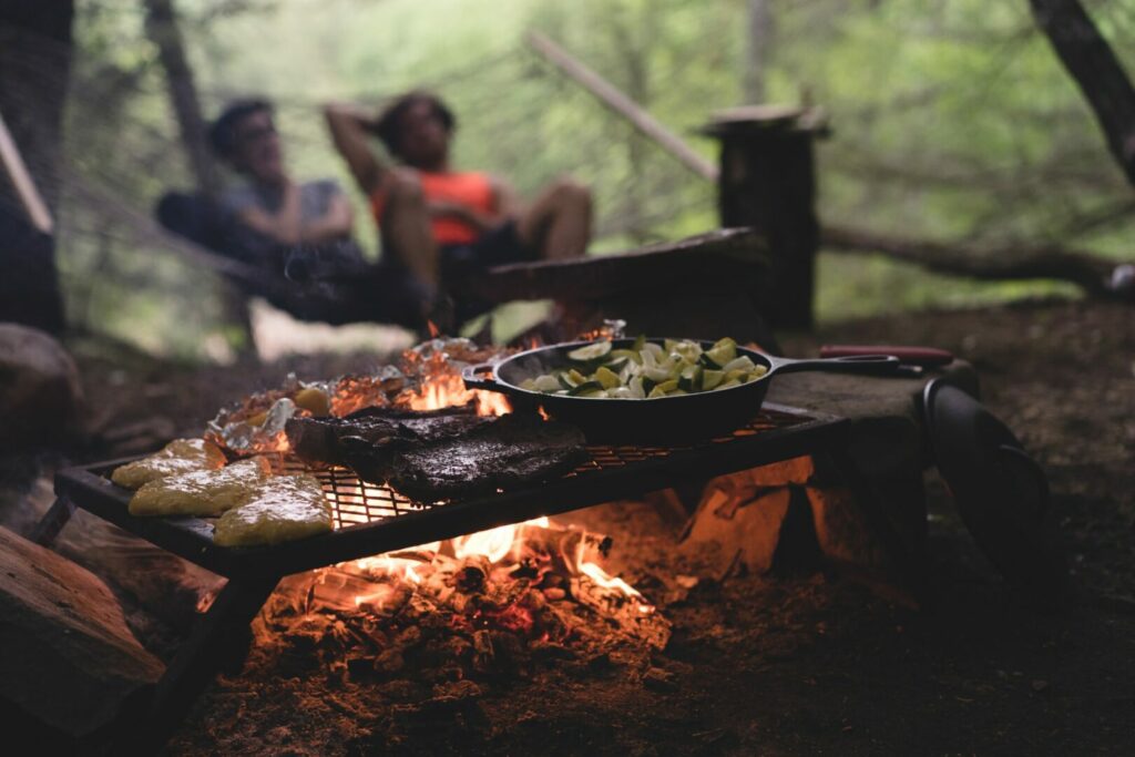 In the foreground, a campfire dinner of chicken, steak, and vegetables in an iron skillet cooks while two campers relax in a hammock in the background.
