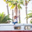 A middle aged woman sit on the top of her camper van with her arms up in excitement in front of a grove of palms trees.