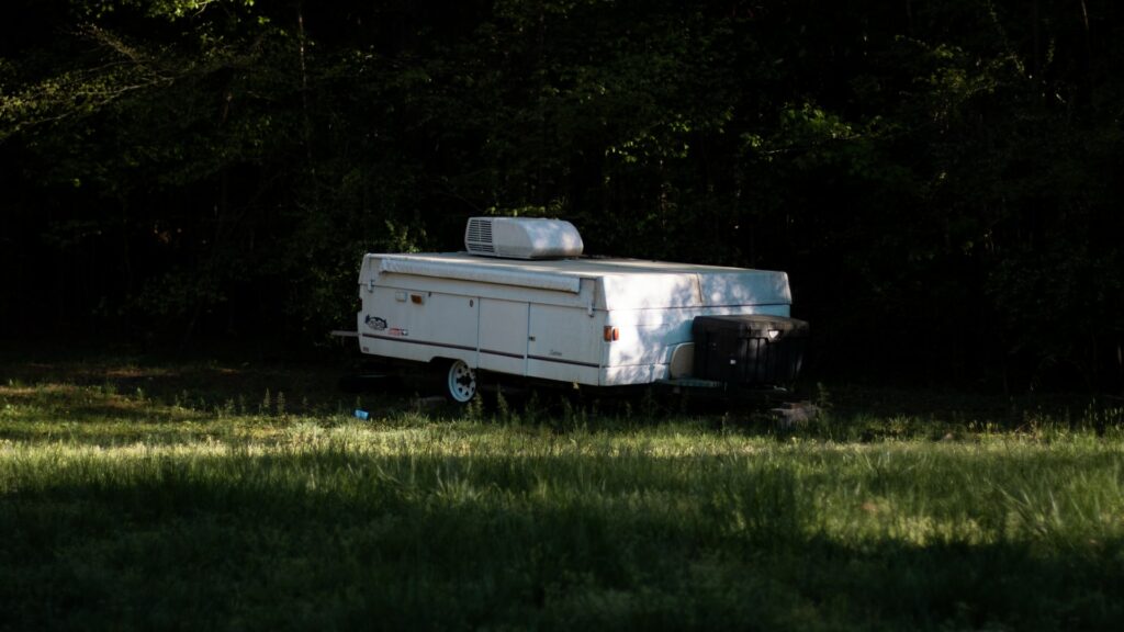 A forgotten pop up camper closed and parked in the shadows next to some trees.