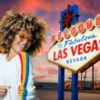 An ethusiastic woman with colored glasses and afro grooves next to a sign welcoming you to Las Vegas in front of an RV campground at sunset