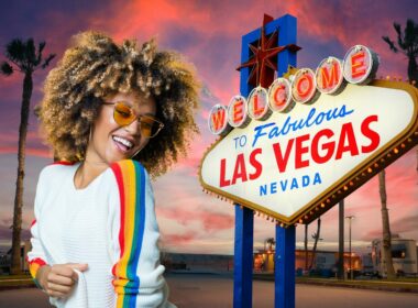 An ethusiastic woman with colored glasses and afro grooves next to a sign welcoming you to Las Vegas in front of an RV campground at sunset