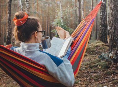 Woman lying in a hammock in a forest drinking coffee reading a book reading as she free camps