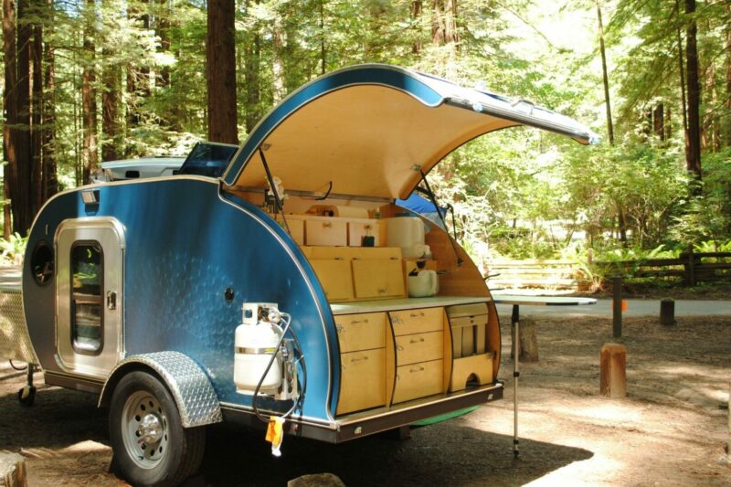 A blue teardrop trailer with a kitchen revealed by opening the back hatch in a wooded campground.