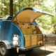 A blue teardrop trailer with a kitchen revealed by opening the back hatch in a wooded campground.