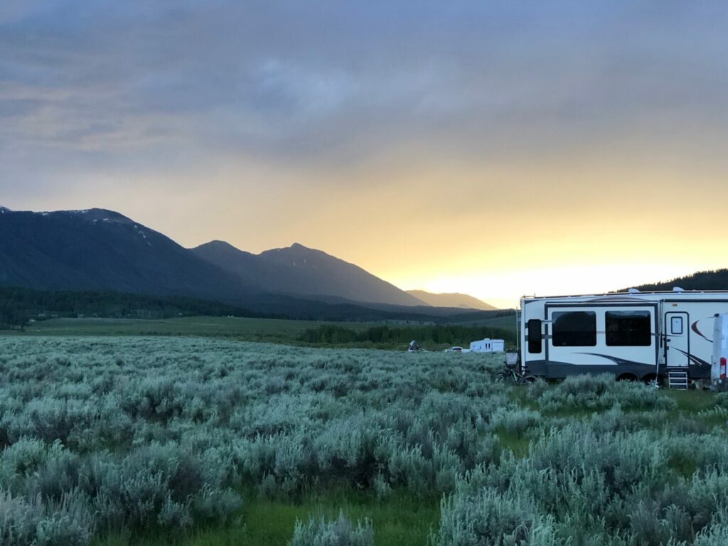 A large RV is boondocking out in an open field with the sun setting behind the mountains.