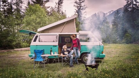 A couple sit outside their camper van with a fire going and mountains in the background.