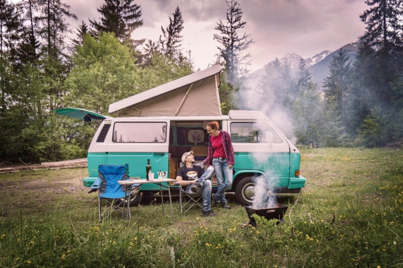A couple sit outside their camper van with a fire going and mountains in the background.