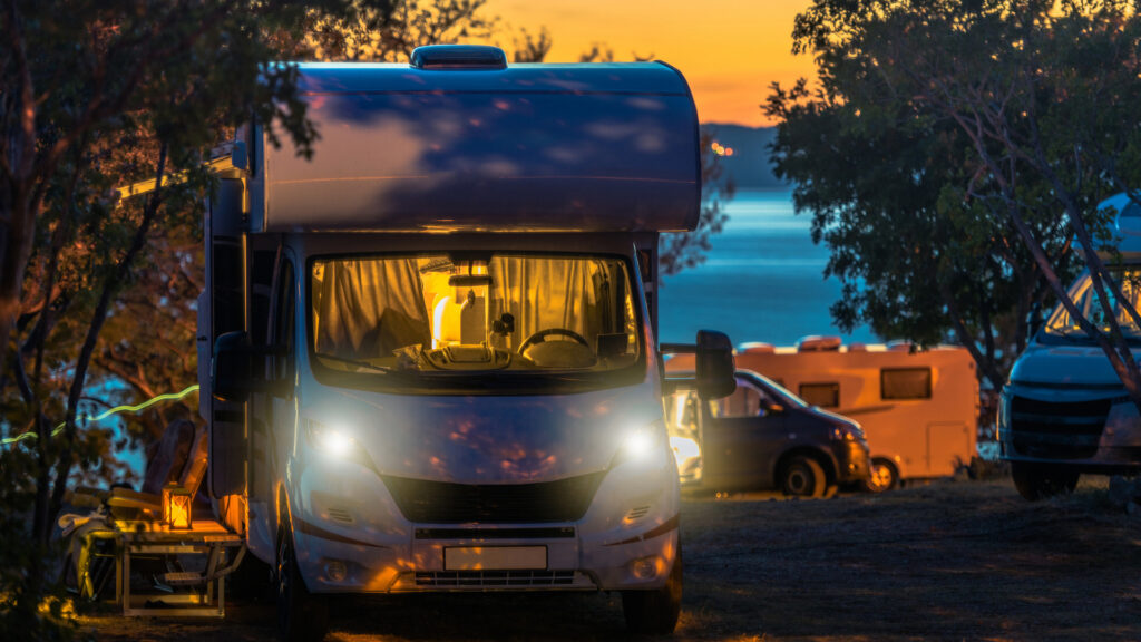 An RV sits in a campground at sunset and is using its headlights and indoor lights to stay bright and visible.