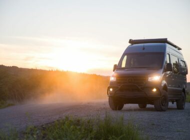 A new luxury off-road camper van on a dirt road as the sunsets and it shines headlights forward.
