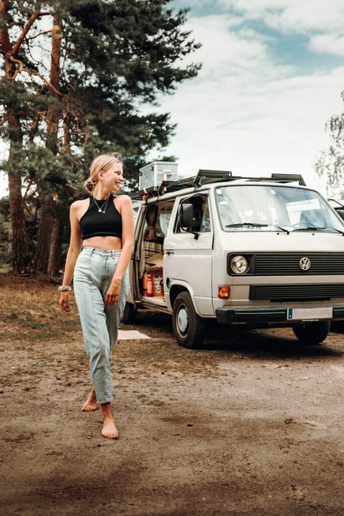 A young woman smiles and walks barefoot outside around her VW camper van parked in the forest