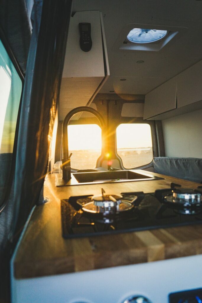 Inside a modern camp van with butcher block counter tops and a residential-style sink as the light from the sunset gleams through the back windows.