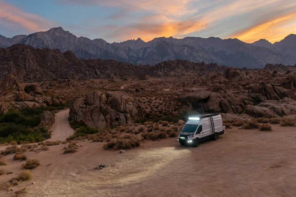 A camper van boondocking as the sun sets over mountains in the desert.