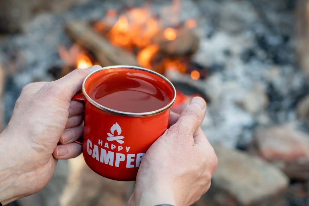 Close up of hands holding a red mug that reads "Happy Camper" while sitting next to a campfire