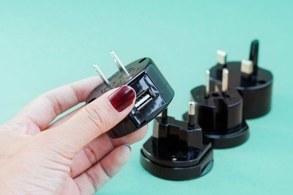 A hand holds up an electrical adapter plug in the foreground with several different types in the background.