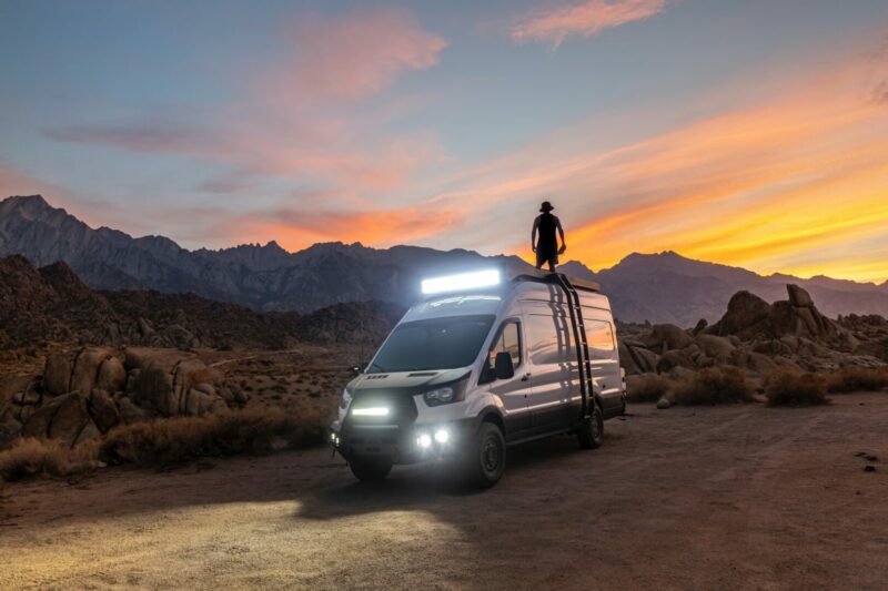 A camper van with electric lights shining through the dark light of dusk as a man stands on top of the van.